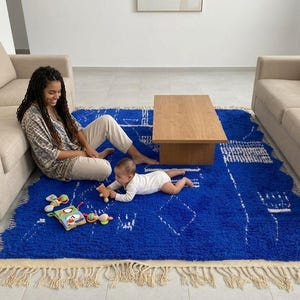 Op de afbeelding: Een helderblauwe vloerkleed met een geometrisch patroon en crèmekleurige franjes. Een vrouw en een baby zitten op het kleed, met een houten salontafel en twee beige banken op de achtergrond. Er liggen speelgoed op het kleed.
