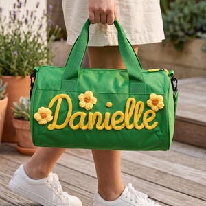 以下が含まれることがあります： 黄色の鉤針編み文字「Danielle」と3つの黄色の花のアクセントが付いた緑色のダッフルバッグ。バッグには緑色のハンドルと黄色のジッパーが付いています。白いスニーカーを履いた人が持っています。