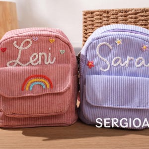Puede incluir: Se muestran dos mochilas de pana, una rosa y otra lavanda. La mochila rosa tiene el nombre "Leni" bordado en blanco, un arcoíris y detalles de corazón. La mochila lavanda presenta el nombre "Sara" y detalles de flores. El texto "SERGIOALE" está en la parte inferior.