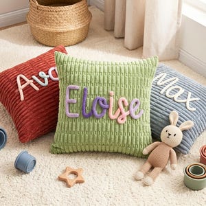 Puede incluir: Tres cojines cuadrados de pana en color óxido, verde salvia y azul pizarra. Los cojines presentan nombres personalizados en letras de hilo en relieve y multicolor: Ava, Eloise y Max. Un pequeño conejo de ganchillo y vasos apilables están cerca.