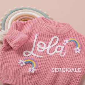 Puede incluir: Jersey de punto rosa con el nombre "Lola" en escritura blanca. El jersey presenta arcoíris y flores bordados. El texto "SERGIOALE" está impreso en blanco. Un adorno de pared con forma de arcoíris está en el fondo.