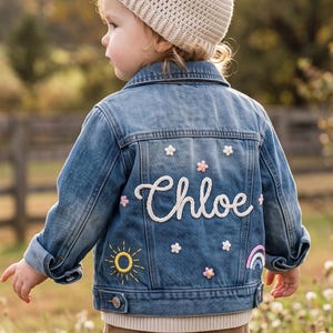 Puede incluir: Una chaqueta de mezclilla azul con el nombre Chloe bordado en letra cursiva blanca en la parte posterior. La chaqueta está decorada con flores bordadas blancas y rosas, un sol amarillo y un pequeño arcoíris. Tiene cuello, mangas largas y botones.