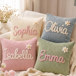 Puede incluir: Cuatro cojines cuadrados de pana en color crema, azul, rosa y verde. Cada cojín presenta un nombre bordado en relieve en letra cursiva: Sophia, Olivia, Isabella y Emma. Una pequeña margarita blanca en tres dimensiones adorna cada nombre.