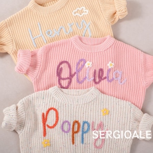 Puede incluir: Tres suéteres de punto en colores pastel con nombres bordados: "Henry" en azul claro, "Olivia" en morado y "Poppy" en colores arcoíris. Cada suéter presenta cuello redondo y mangas cortas, con detalles florales.