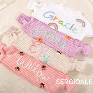 Puede incluir: Cuatro suéteres de punto de colores pastel con nombres bordados y elementos decorativos. Los suéteres son blancos, lavanda, beige y rosa. Los nombres "Gracie", "Sahra", "Elly" y "Willow" están bordados en cursiva. Los suéteres tienen puños y cuello acanalados.