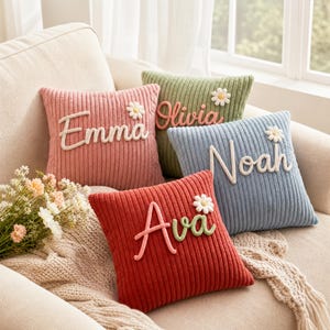 Puede incluir: Cuatro cojines cuadrados de pana en rosa, verde, azul y rojo. Cada cojín presenta un nombre en relieve escrito en cursiva con hilo blanco o verde y una pequeña aplicación de margarita blanca. Los nombres mostrados son Emma, Olivia, Noah y Ava.