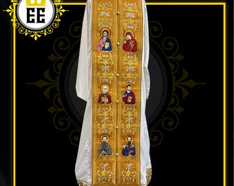 Pode incluir: Uma vestimenta litúrgica branca com bordados e franjas douradas. A vestimenta apresenta figuras religiosas bordadas com detalhes coloridos. O bordado dourado inclui padrões florais e geométricos. A vestimenta é exibida em um manequim.