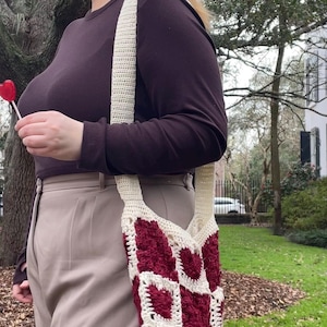 Peut inclure: Un sac fourre-tout en crochet fait à la main, avec une bandoulière et un corps de couleur crème. Le sac présente un motif de cœurs bordeaux. La personne porte une chemise à manches longues bordeaux et un pantalon beige. Une sucette rouge en forme de cœur est tenue.