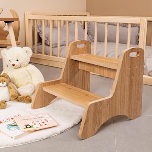 May include: A two-step wooden step stool with a natural finish. The stool has a handle cut-out on each side and is designed for children. A children's book and stuffed animals are in the background.