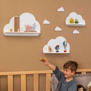 May include: White cloud-shaped shelves with various miniature figurines and houses. The shelves are mounted on a brown wall. A child is holding a toy airplane in front of the shelves. The shelves are a decorative element for a child's room.