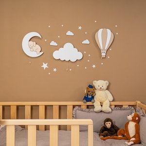 Wooden nursery wall decor set with elephant on moon, cloud and hot air balloon night light above natural pine baby crib, daytime view without light, star wall stickers on warm brown nursery wall, boho gender neutral baby room wall art, handmade plywood moon cloud balloon wall hanging, montessori style toddler bedroom decoration, minimalist scandinavian nursery decor, newborn baby shower gift set idea