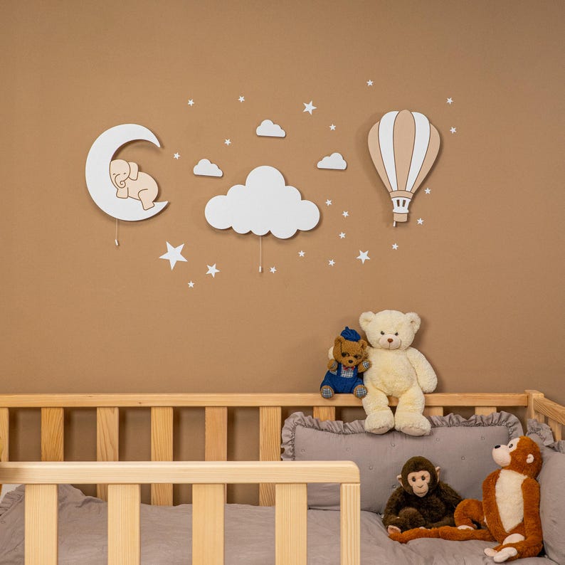Wooden nursery wall decor set with elephant on moon, cloud and hot air balloon night light above natural pine baby crib, daytime view without light, star wall stickers on warm brown nursery wall, boho gender neutral baby room wall art, handmade plywood moon cloud balloon wall hanging, montessori style toddler bedroom decoration, minimalist scandinavian nursery decor, newborn baby shower gift set idea