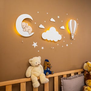 Full nursery night light set glowing above natural pine baby crib, wooden elephant on moon, large cloud and hot air balloon wall lamps with warm LED backlight, star stickers on brown wall, stuffed animals in crib, cozy nighttime nursery atmosphere, boho gender neutral baby room wall art, handmade plywood moon cloud balloon lamp set, montessori style toddler bedroom, newborn baby shower gift idea