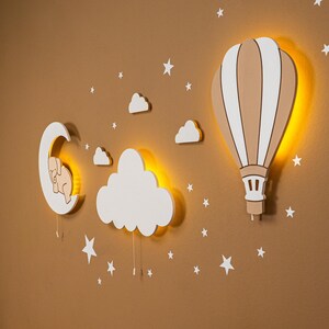 Glowing hot air balloon and cloud wooden night light on warm brown nursery wall, elephant moon wall decor with star stickers above pine baby crib, warm LED backlight detail, close up side angle view of nursery night light set, boho baby room wall art, handmade plywood balloon lamp for kids bedroom, gender neutral toddler room decoration, montessori nursery wall accessory, baby shower gift, scandinavian style children room night light