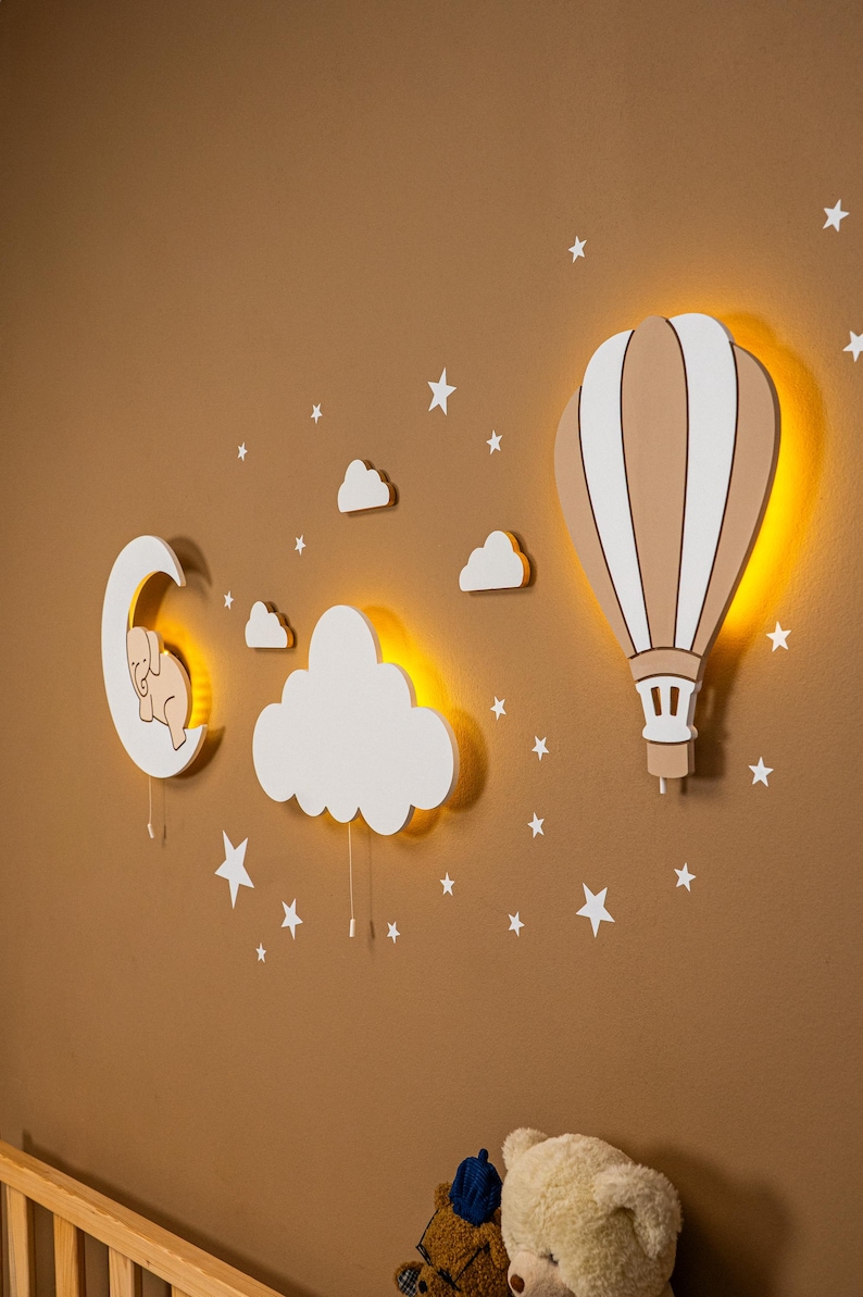 Glowing hot air balloon and cloud wooden night light on warm brown nursery wall, elephant moon wall decor with star stickers above pine baby crib, warm LED backlight detail, close up side angle view of nursery night light set, boho baby room wall art, handmade plywood balloon lamp for kids bedroom, gender neutral toddler room decoration, montessori nursery wall accessory, baby shower gift, scandinavian style children room night light