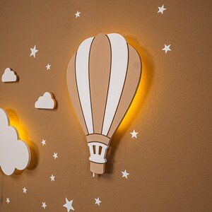 Close up of wooden hot air balloon night light with warm LED backlight glow, handmade plywood balloon wall lamp detail, white cloud and elephant moon decor partially visible, star stickers on warm brown nursery wall, natural wood texture and painted stripe detail on balloon lamp, boho nursery wall art, gender neutral baby room night light, montessori style kids bedroom wall decor, toddler room lamp, baby shower gift