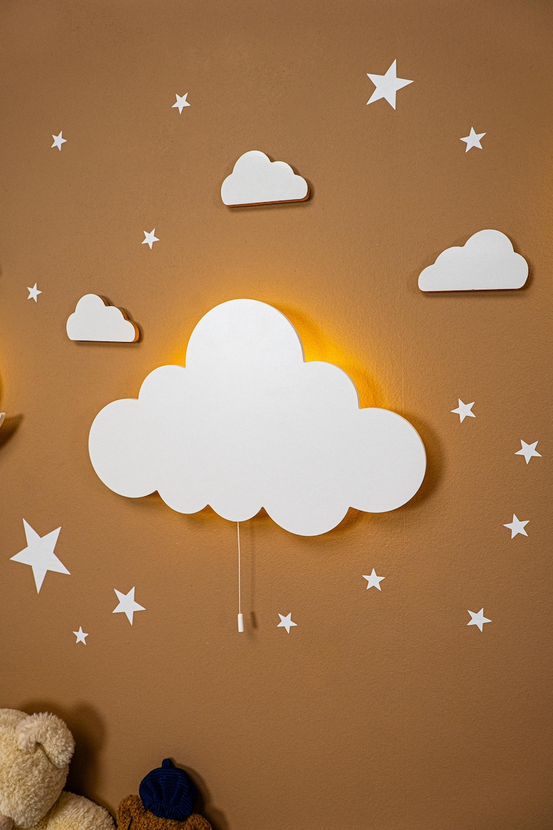 Cloud-shaped wooden night light for baby nursery wall decor, warm LED cloud lamp with hanging stars and clouds, Montessori inspired nursery lighting, neutral beige kids room decoration, handcrafted plywood night light for toddler bedroom, soft ambient nursery lamp for baby room, modern Scandinavian style wall light for children’s room.
