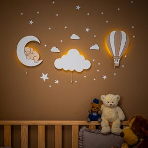 May include: A nursery wall decor set featuring a crescent moon with an elephant, clouds, a hot air balloon, and stars. The white wooden cutouts have warm yellow lighting. A wooden crib and teddy bears are in the foreground.