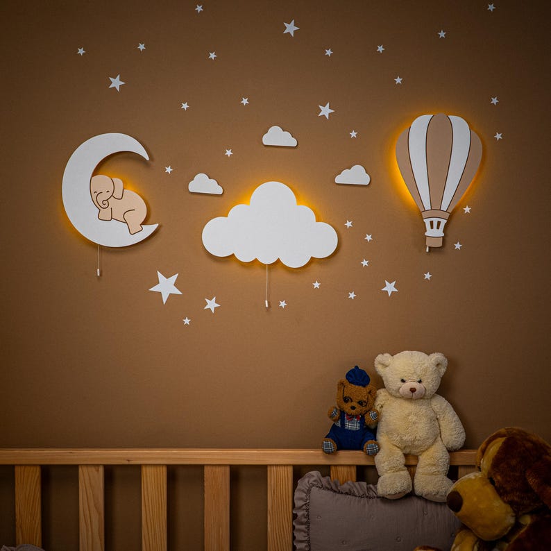 Wooden nursery night light set with elephant on moon, cloud and hot air balloon wall decor, warm LED backlight glow above natural pine baby crib, star wall stickers on brown nursery wall, boho gender neutral baby room wall art, handmade plywood moon cloud balloon lamp set, montessori style toddler bedroom decoration, newborn baby shower gift idea, scandinavian kids room night light