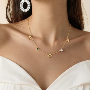 Puede incluir: Collar MOM dorado con una pequeña gema verde y una gema transparente. El collar tiene las letras M, O y M. Un pendiente grande, redondo, de color plateado con piedras transparentes es visible en el fondo.