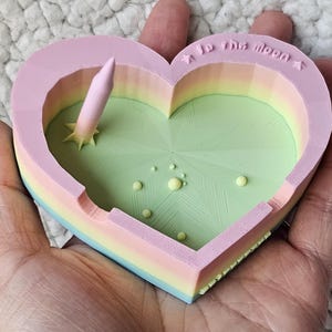 May include: A heart-shaped trinket dish in pastel colors, with a rocket-like structure and the text "to the moon". The dish features a light green interior with small yellow spheres, and a multi-colored exterior.