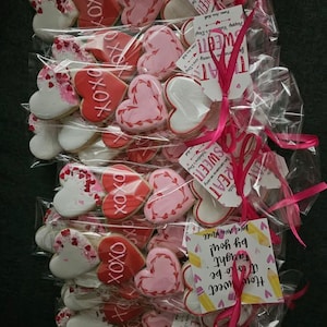May include: Arrangement of heart-shaped cookies in clear cellophane bags, tied with pink ribbons. Cookies feature white, pink, and red icing with decorative details and the text "XOXO". Tags with text are attached.