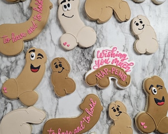Op de afbeelding: Een verzameling fallusvormige koekjes in verschillende tinten beige en bruin. Sommige koekjes hebben cartoongezichtjes, terwijl andere roze glazuurteksten bevatten zoals "To have and to hold" of "Wishing you much HAP-PENIS" in cursief schrift.