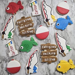 May include: A collection of decorated sugar cookies with a fishing theme. The cookies include red, blue, green, and yellow fish, bobbers, fishing poles, and signs that say "Fishing Hole", "Boat Ramp", and "Bait Shop". Some cookies are shaped like the number one with the name "Carter".