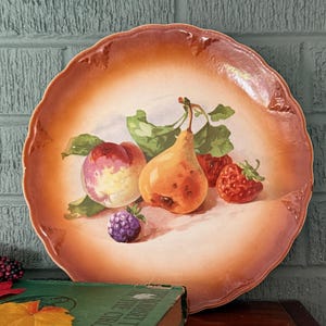 May include: A decorative plate with a painted still life of fruit. The plate features a peach, pear, strawberries, and a blackberry. The plate has a scalloped edge with a brown and cream color scheme.
