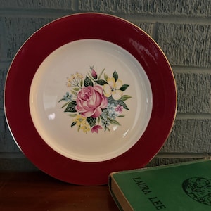 May include: A round, vintage-style plate with a maroon rim and a white center. The center features a floral bouquet with pink roses, yellow and blue flowers. The plate has a gold trim. A green book is in the foreground.