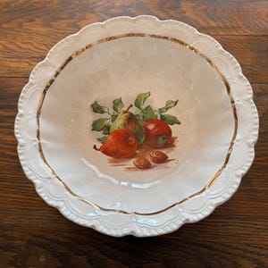 May include: White ceramic serving bowl with a scalloped edge and gold trim. The bowl features a painted still life of pears, apples, and almonds. The fruit is depicted in shades of red, green, and brown.