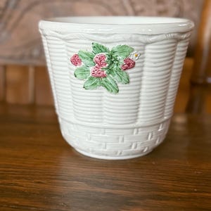 May include: White ceramic planter with a basket weave design. The planter features a raised floral design with pink strawberries and green leaves. The planter is suitable for indoor or outdoor use.