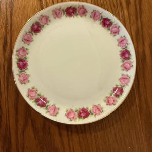 May include: A round, off-white plate with a gold rim. The plate is decorated with a ring of pink and red roses around the edge. The plate is sitting on a wooden surface.