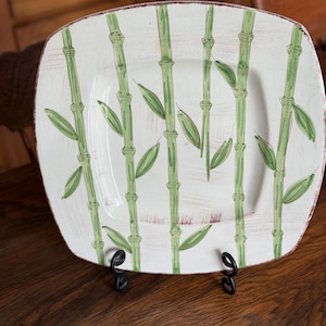May include: Square ceramic plate with a bamboo design. The plate features a white background with green bamboo stalks and leaves. The edges have a reddish-brown trim. The plate is displayed on a black metal stand.