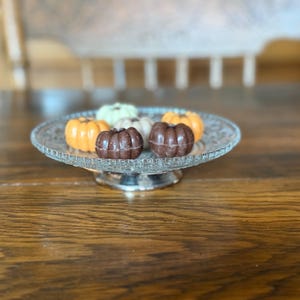 May include: A decorative glass serving plate with a silver base holds six small, pumpkin-shaped objects. The pumpkins are in shades of orange, brown, and cream. The plate is on a wooden surface.
