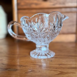 May include: A clear, cut-glass creamer with a handle and a spout. The creamer has a scalloped rim and a pedestal base. The glass is intricately patterned with a starburst design. The creamer is sitting on a wooden surface.