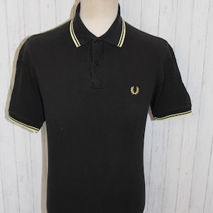 Könnte beinhalten: Schwarzes Poloshirt mit gelbem Kragen und Ärmelbesatz. Das Shirt hat eine Knopfleiste mit drei Knöpfen und ein kleines gesticktes Emblem auf der Brust. Das Kleidungsstück wird auf einer Schaufensterpuppe vor einem weißen Holzhintergrund präsentiert.