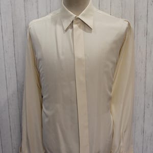 以下が含まれることがあります： クリーム色の長袖ドレスシャツ。クラシックカラーとボタン付きカフスが特徴です。 シャツの前面には縦のストライプがあり、マネキンに着せられています。 生地は滑らかで、わずかな光沢があります。