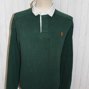 May include: A dark green long-sleeve rugby shirt with a white collar and a three-button placket. An orange logo is on the chest. The shirt is displayed on a mannequin against a white wood-paneled background.