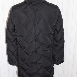 Puede incluir: Abrigo negro acolchado con cuello alto y cierre de botones. El abrigo tiene un patrón de diamantes y mangas largas. El abrigo se exhibe en un maniquí. Un abrigo de invierno.