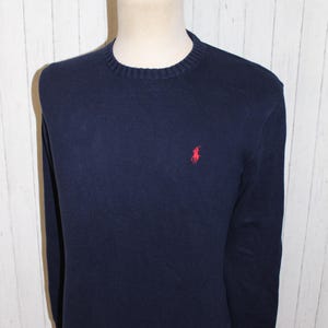 May include: A navy blue long-sleeve sweater with a ribbed crew neckline, cuffs, and hem. The garment features a small, embroidered red logo of a person on a horse playing polo, positioned on the left side of the chest against the dark fabric.
