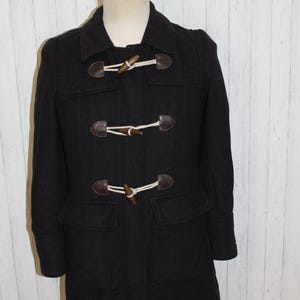 May include: A black duffel coat with a collared neckline and two front pockets. The coat features wooden toggle closures with leather accents. The coat is displayed on a mannequin against a white and wood-paneled background.