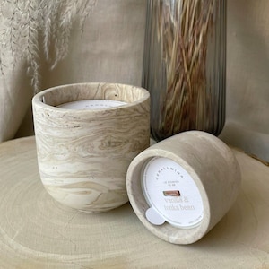 Könnte beinhalten: Zwei beige und cremefarbene marmorierte Kerzen, eine aufrecht und eine auf der Seite, mit einem Etikett mit der Aufschrift "vanilla & tonka bean". Eine klare Glasvase mit getrocknetem Ziergras befindet sich im Hintergrund. Die Kerzen stehen auf einer Holzoberfläche.
