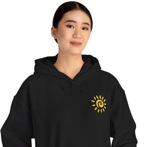 Può includere: Felpa con cappuccio nera con una grafica a forma di sole giallo sulla parte superiore sinistra del petto. La felpa ha una tasca frontale e un cappuccio con coulisse. La modella indossa pantaloni neri.