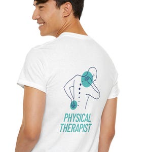Puede incluir: Camiseta blanca con un gráfico turquesa y azul marino de una persona con puntos de dolor en la espalda y el cuello. El texto "PHYSICAL THERAPIST" está debajo del gráfico. La persona lleva vaqueros azules.