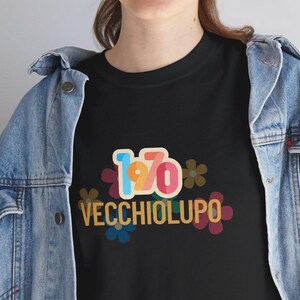 Può includere: T-shirt nera con un design retrò con il numero "1970" in blu, giallo e rosso, e la parola "VECCHIOLUPO" in un font vintage. Il design include accenti floreali in vari colori.