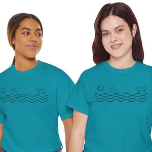 Può includere: Due persone indossano magliette turchesi con un disegno a tratto nero di un sole, onde e creature marine. Le magliette sono a maniche corte e abbinate a jeans blu. L'immagine è una ripresa media.
