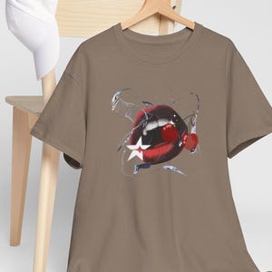 May include: A brown t-shirt with a graphic design of red lips, a white star, and cherries. The t-shirt is hanging on a wooden hanger. A white baseball cap rests on a wooden chair.