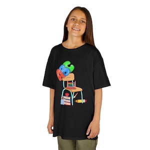 Può includere: T-shirt nera con un'illustrazione colorata di un banco di scuola, un abaco, libri e una matita. Il design include le lettere A, B e C in colori vivaci. La maglietta è indossata da una persona.