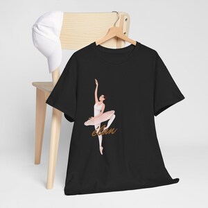 Può includere: T-shirt nero con un'illustrazione di ballerina in body bianco e tutù rosa, con la parola "élan" in oro. La ballerina è in posa aggraziata con un braccio alzato. Un cappello bianco è appoggiato su una sedia di legno.